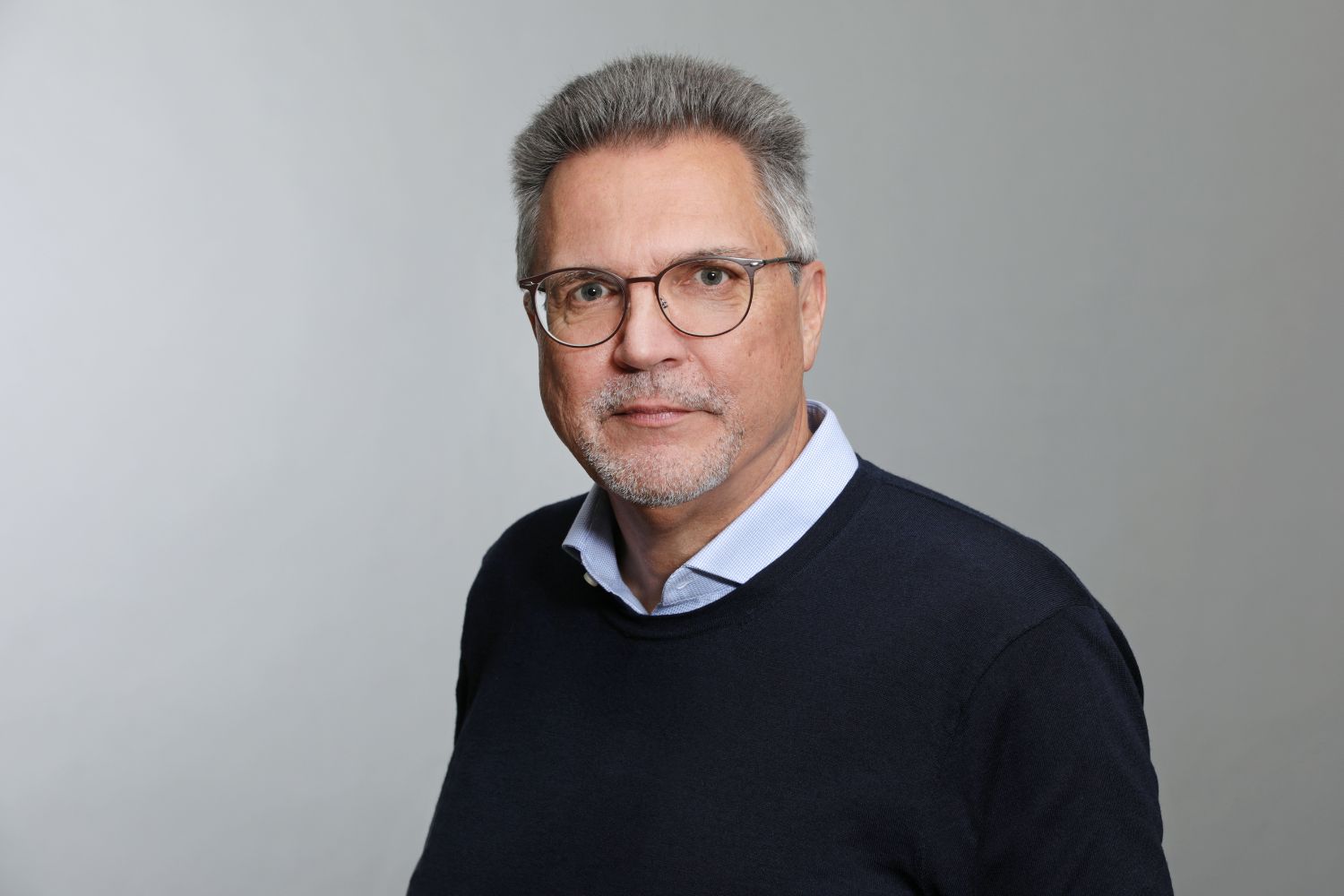 „Porträt eines Mannes mittleren Alters mit grauen Haaren, Brille und kurzem Bart. Er trägt ein dunkelblaues Pullover über einem hellblauen Hemd und blickt freundlich in die Kamera vor neutral grauem Hintergrund.“