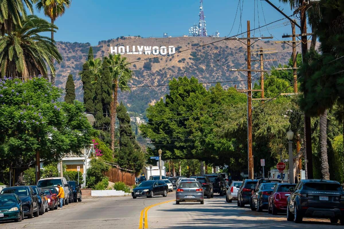 Blick auf eine von Bäumen gesäumte Straße in Los Angeles mit dem berühmten Hollywood-Schriftzug im Hintergrund unter einem klaren, hellblauen Tageshimmel.
