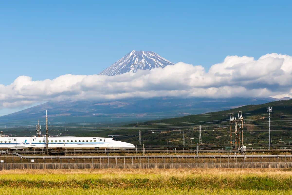 Ein weißer Shinkansen-Schnellzug fährt vor dem schneebedeckten Gipfel des Fuji, der teilweise von einer Wolkenschicht umgeben ist, durch eine ländliche Region.