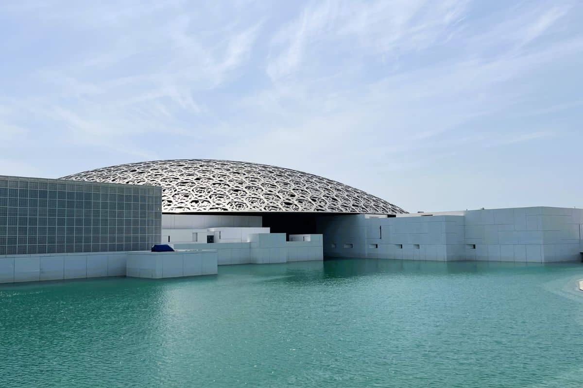Die markante, durchbrochene Kuppel des Louvre Abu Dhabi erhebt sich über einer modernen, weißen Gebäudestruktur, die von einer türkisfarbenen Wasserfläche umgeben ist.