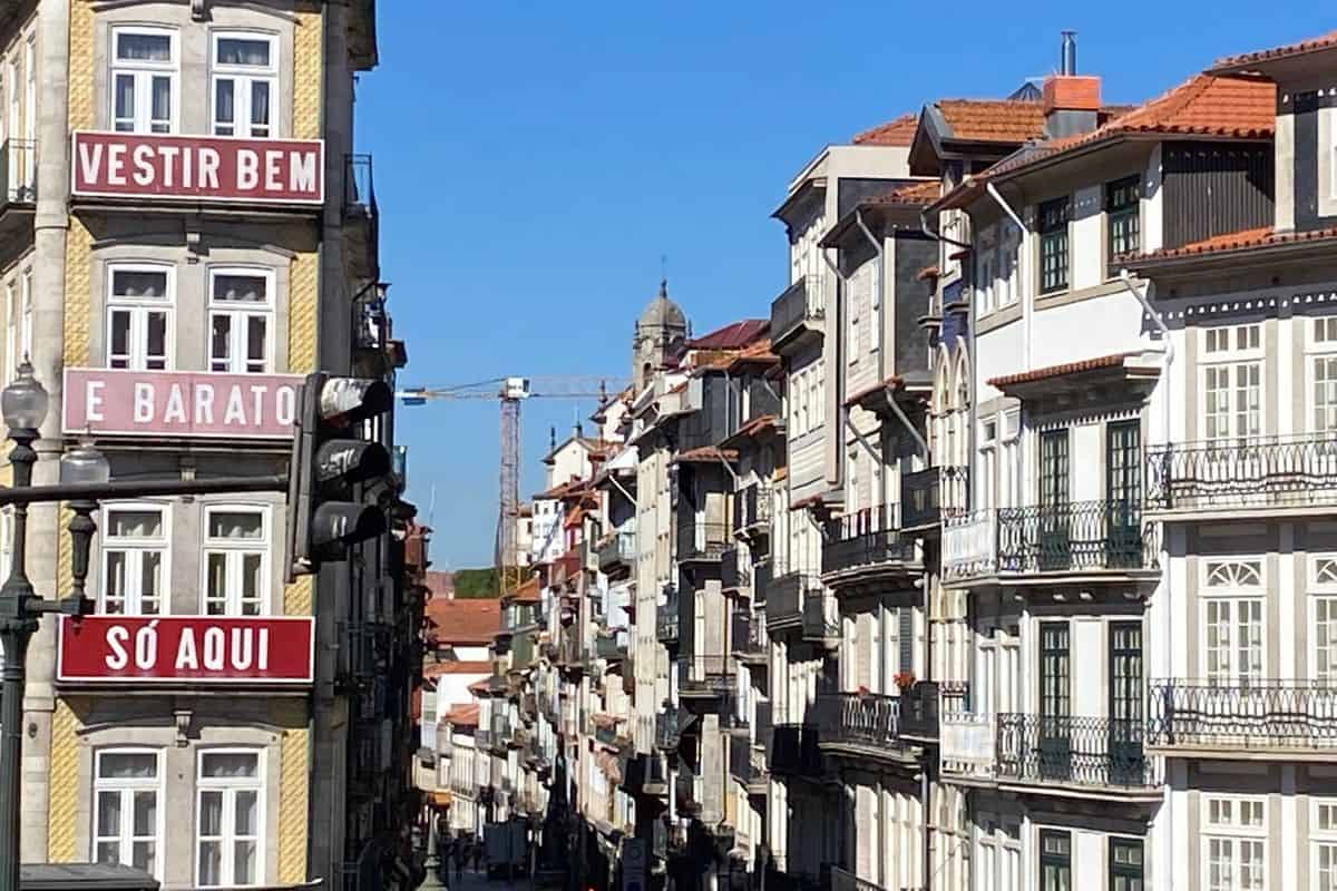 Blick in eine enge Gasse mit historischen Gebäuden, die mit Balkonen und Schildern versehen sind, unter einem klaren blauen Himmel bei hellem Tageslicht.