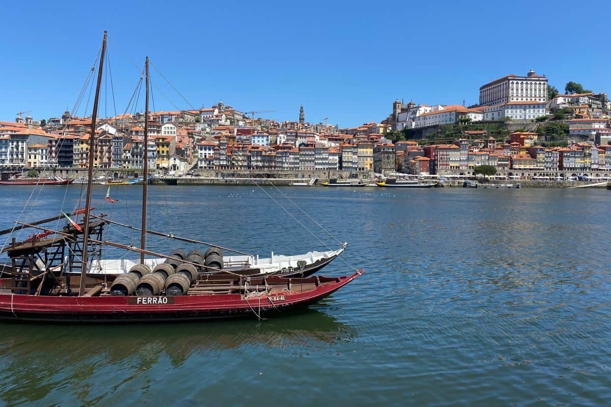 Traditionelle Holzboote mit Weinfässern an Deck liegen im Fluss Douro vor der historischen Kulisse der Stadt Porto unter einem klaren, blauen Sommerhimmel.
