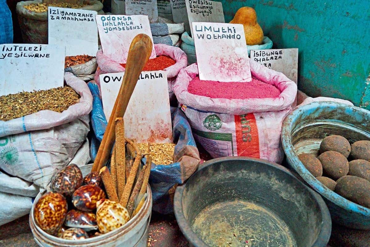 Marktstand mit Säcken voller bunter Pulver, Kräuter und Muscheln in Schalen. Weiße Schilder mit handgeschriebenen Bezeichnungen stecken in den Warenbehältern.