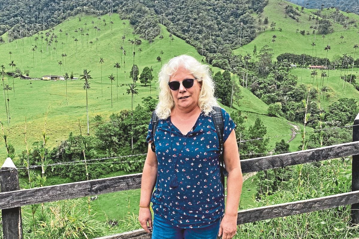 Eine Frau mit blondem Haar und Sonnenbrille steht vor einem Holzzaun in einer hügeligen Landschaft mit zahlreichen hohen Wachspalmen im Hintergrund.