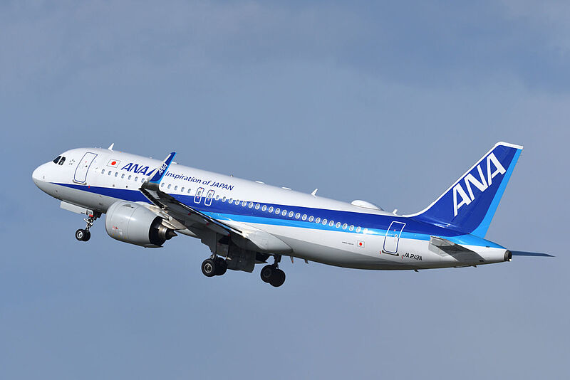 Ein fliegender Airbus A320 der Fluggesellschaft All Nippon Airways (ANA) in weiß-blauer Lackierung vor bewölktem Himmel und von der Sonne angestrahlt.