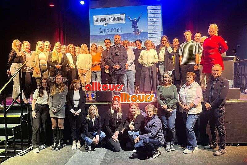 Gruppenfoto von Teilnehmenden der Alltours-Roadshow 2025 auf einer Bühne vor einer Präsentationsleinwand mit dem Alltours-Logo und dem Schriftzug „Alltours Roadshow – Guten Appetit“.