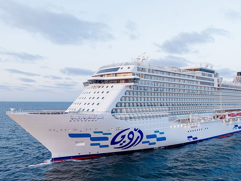 Seitenansicht eines Kreuzfahrtschiffes auf See. Am weißen Rumpf prangt das arabische Reederei-Logo in blauem Design.