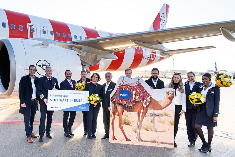 Die Bordcrew eines Condor-Flugzeugs steht mit einem Schild, auf dem „Inaugural Flight Stuttgart – Dubai“ steht, sowie einem Aufsteller, auf dem ein Kamel abgebildet ist, vor einem Flugzeug von Condor auf dem Vorfeld des Flughafens Stuttgart.