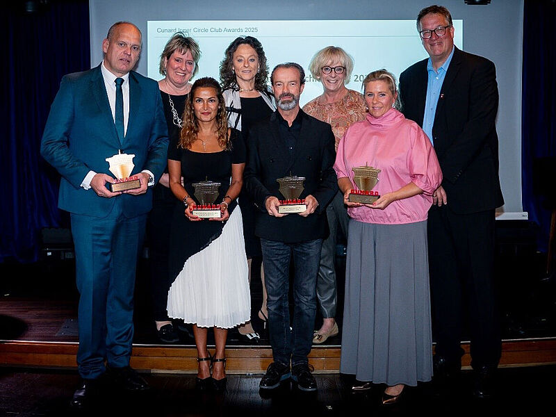 Gruppenfoto: Preisträger der Cunard Inner Circle Club Awards 2025 mit Trophäen auf einer Bühne vor Präsentationsleinwand an Bord des Kreuzfahrtschiffes Queen Anne.