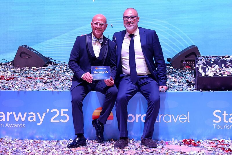 Zwei Männer sitzen auf einer Bühne und lächeln in die Kamera.