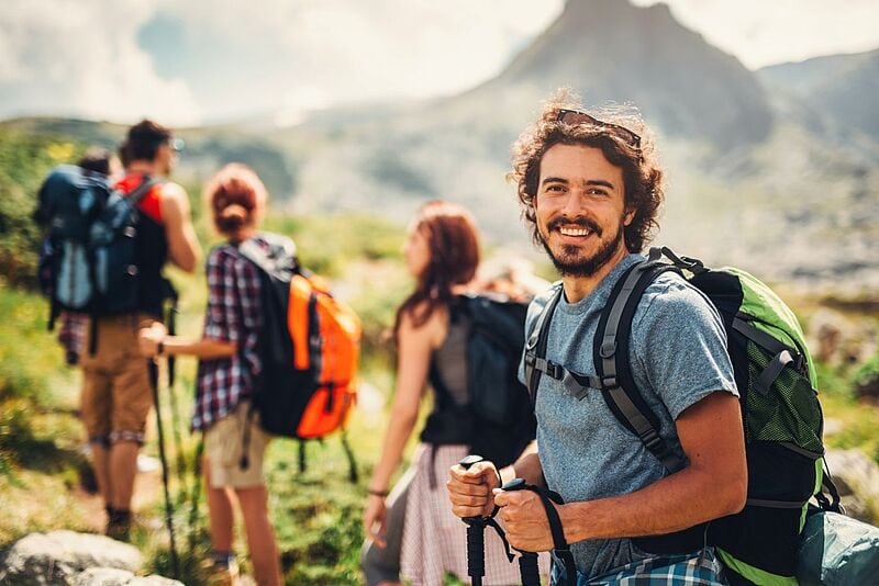 Eine Gruppe von Wandernden auf einem Bergpfad; im Vordergrund steht ein junger Mann mit Rucksack und Trekkingstöcken