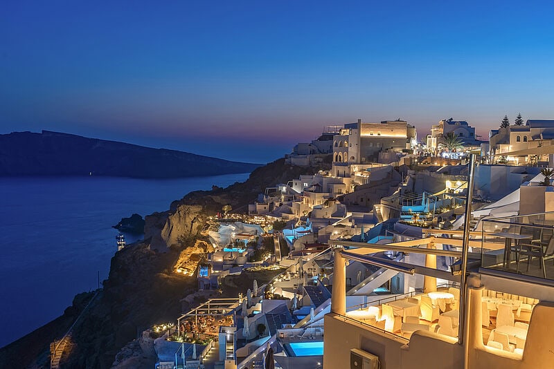Weiße Häuser im Küstenstädtchen Oia auf der Insel Santorin an einem steilen Berghang, dazwischen blau beleuchtete Pools, blaue Stundezwischen 