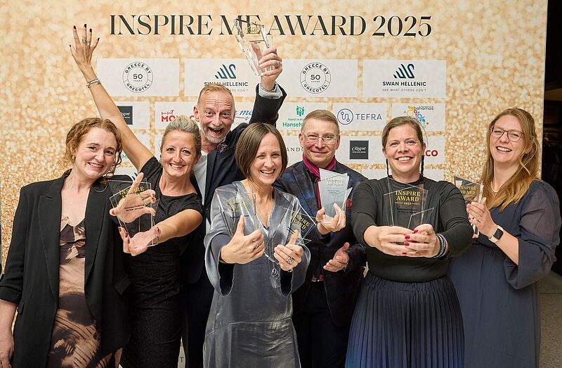 Luxusurlaub, Kiticon, Silvio Rebmann, Cuba Eine Gruppe von sieben fröhlichen Personen posiert vor einer goldenen Fotowand mit der Aufschrift „INSPIRE ME AWARD 2025“. Alle halten gläserne Trophäen in den Händen und lächeln in die Kamera. Im Hintergrund sind Logos verschiedener Sponsoren zu sehen.