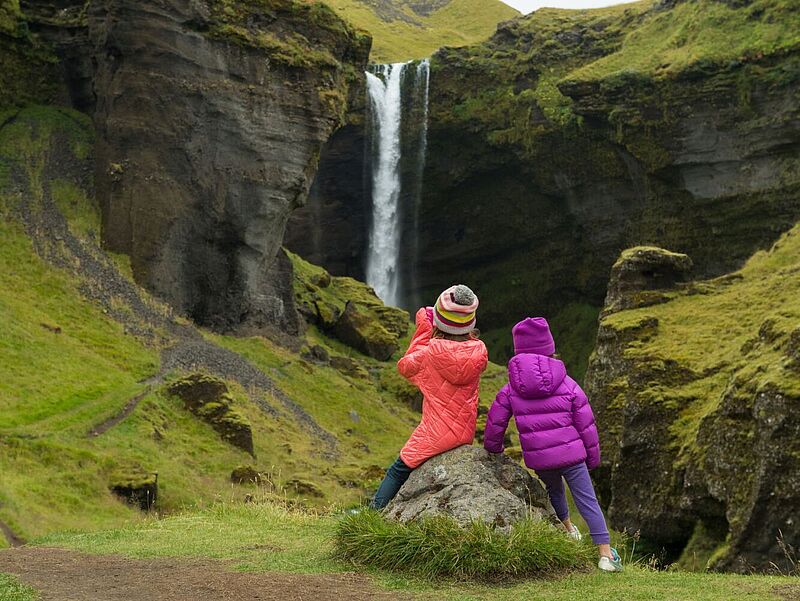 Zwei Mädchen in Rückansicht mit Mützen und Winterjacken schauen auf einen Wasserfall, der von grün bewachsenen Felsen herunterstürzt.