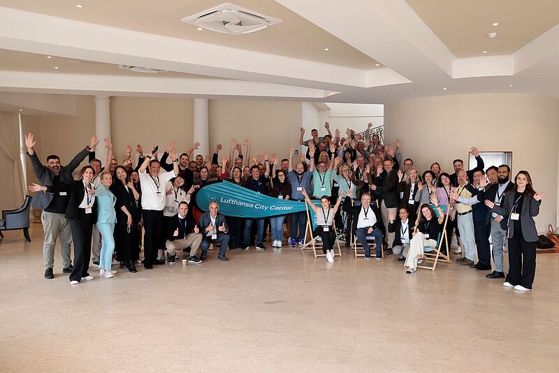 Eine große Gruppe von Teilnehmerinnen und Teilnehmern der Lufthansa City Center Vollversammlung posiert in einem hellen Konferenzraum. Alle lächeln, heben die Arme zum Gruß und stehen oder sitzen in mehreren Reihen. In der Mitte halten zwei Personen ein türkisfarbenes Banner mit der Aufschrift „Lufthansa City Center“.