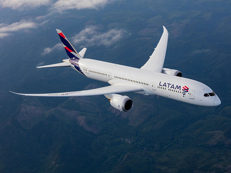 Draufsicht auf ein Flugzeug der Airline Latam in weiß-blau-roter Lackierung, das über eine grüne Gebirgslandschaft fliegt. Vereinzelt sind Wolken zu sehen.