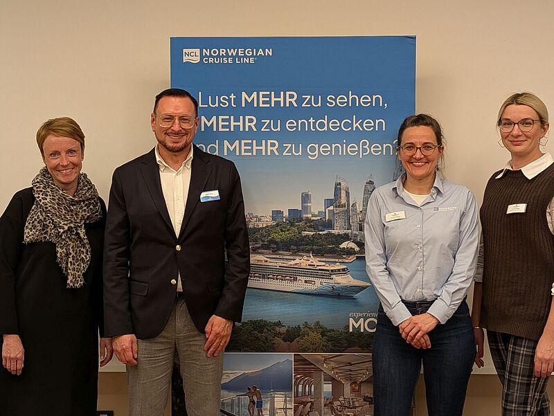 Vier Personen stehen lächelnd vor einem blauen Banner von Norwegian Cruise Line mit dem Schriftzug „Lust MEHR zu sehen, MEHR zu entdecken und MEHR zu genießen“. Die Gruppe besteht aus drei Frauen und einem Mann. Alle tragen Namensschilder und sind geschäftlich gekleidet. Das Banner zeigt im Hintergrund eine Skyline, ein Kreuzfahrtschiff und eine Küstenlandschaft.