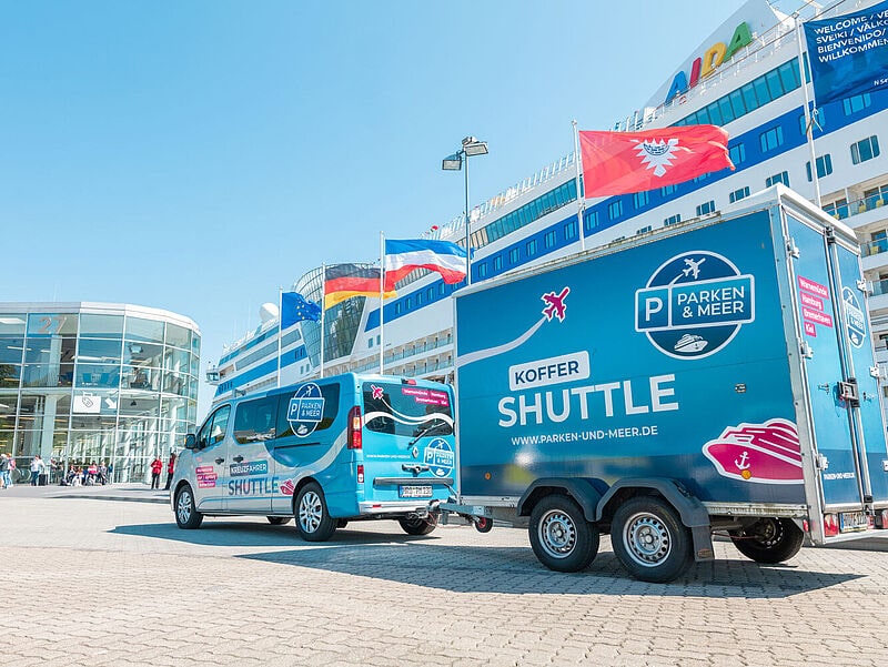 Blauer Shuttle-Van mit Anhänger vor Kreuzfahrtterminal, Logo „Parken & Meer Koffer Shuttle“ auf Fahrzeugen, Kreuzfahrtschiff im Hintergrund.