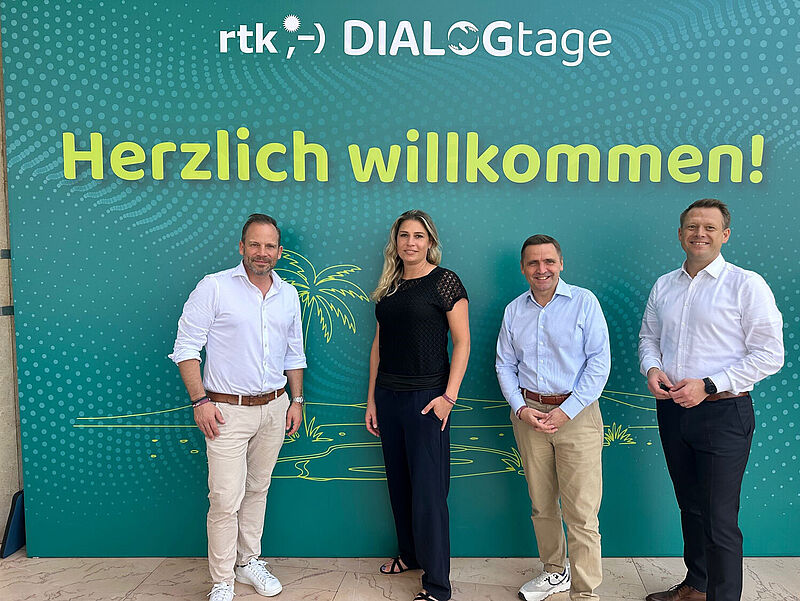 Vier Touristiker stehen vor einer Fotowand. Die Frau und die drei Männer schauen lächelnd in die Kamera. 