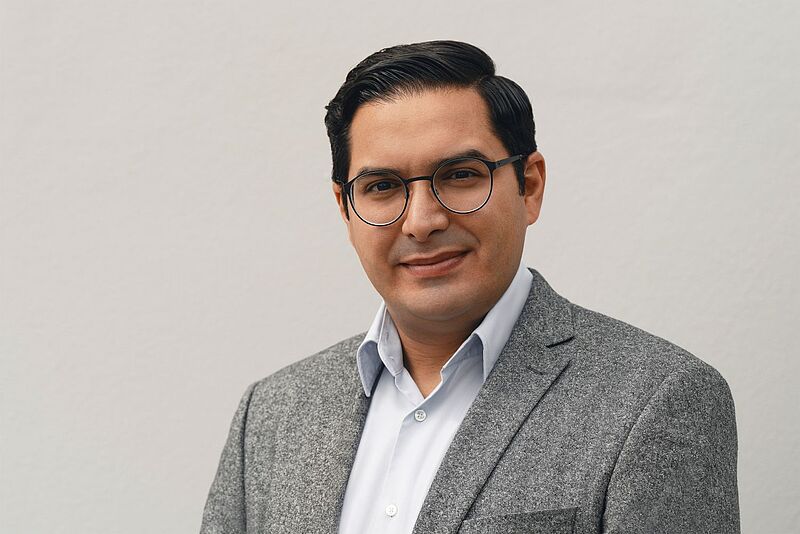 Porträt eines Mannes mit dunklem, seitlich gekämmtem Haar und runder Brille. Er trägt ein graues Sakko über einem hellen Hemd und blickt freundlich in die Kamera; neutraler, heller Hintergrund.