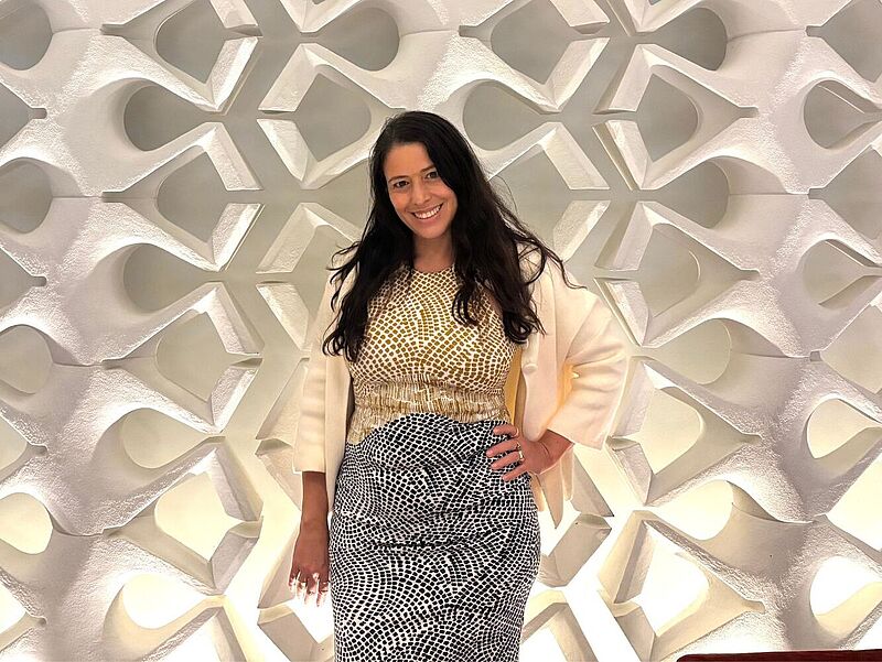 Eine Frau steht vor einer dekorativen, beleuchteten Wand mit geometrischem Muster. Sie trägt ein gelb-weiß gemustertes Oberteil, einen cremefarbenen Blazer und einen schwarz-weiß gemusterten Rock. 