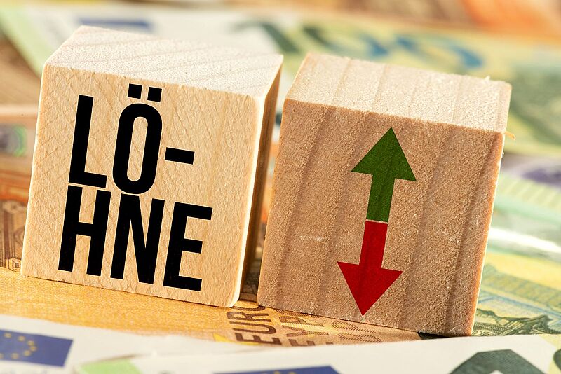Holzklötze mit dem Wort „Löhne“ und einem Symbol aus grünem Aufwärtspfeil und rotem Abwärtspfeil, im Hintergrund liegen unscharf Euro-Geldscheine – Symbolbild für Lohnverhandlungen oder Tarifauseinandersetzungen.