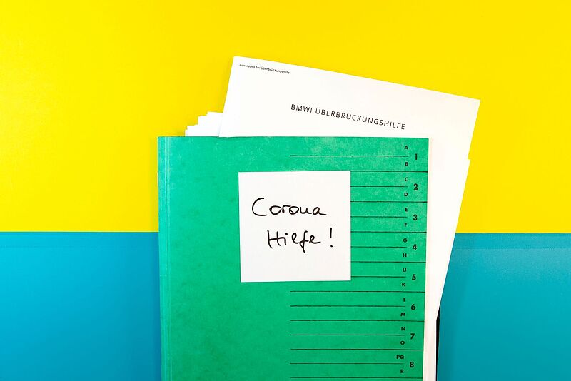 Ein grüner Aktenordner liegt vor gelbem und blauem Hintergrund, darauf klebt ein Zettel mit der Aufschrift „Corona Hilfe“, und oben ragt ein Formular mit dem Titel „BMWi Überbrückungshilfe“ heraus – Symbolbild für staatliche Coronahilfen und Überbrückungshilfe während der Pandemie.