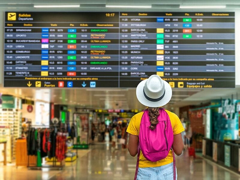 Eine Person mit weißem Hut, gelbem T-Shirt und pinkem Rucksack steht in einem Flughafen-Terminal und blickt auf eine große Anzeigetafel mit Abflügen. Im Hintergrund sind Geschäfte und Reisende zu sehen.