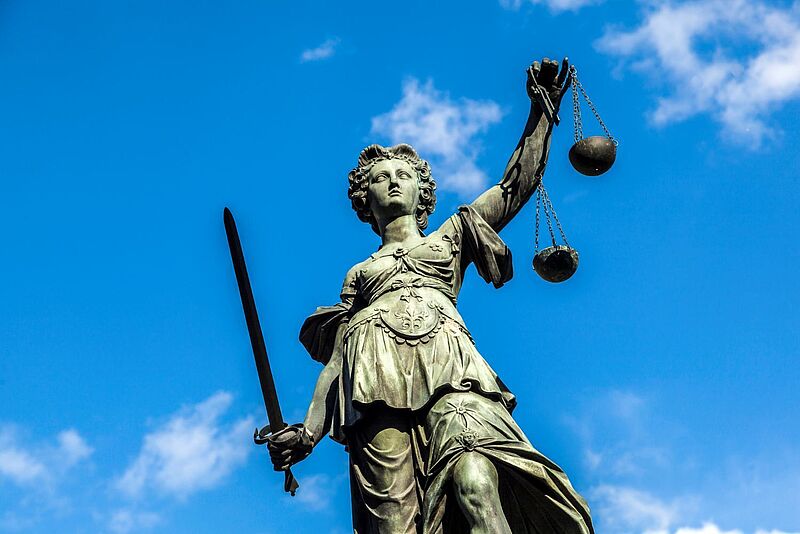 Bronzene Justitia-Statue vor blauem Himmel, mit erhobenem Schwert in der rechten Hand und einer Waage in der linken Hand.