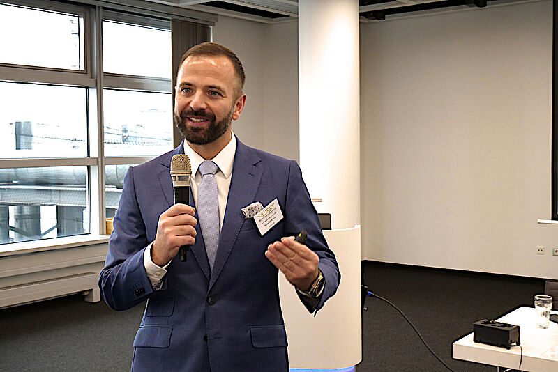 Ein jüngerer Mann im grauen Anzug, weißem Hemd und Krawatte hält mit einem Mikrofon in der Hand einen Vortrag in einem Konferenzraum und gestikuliert