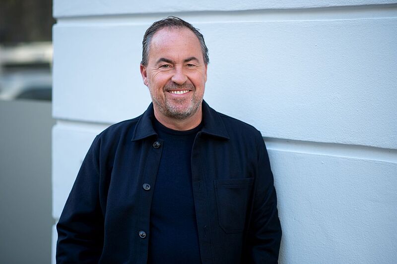 Ein lächelnder Mann mittleren Alters mit kurzem Haar und Bart steht lässig an einer weißen Wand. Er trägt eine dunkelblaue Jacke und ein passendes Oberteil.