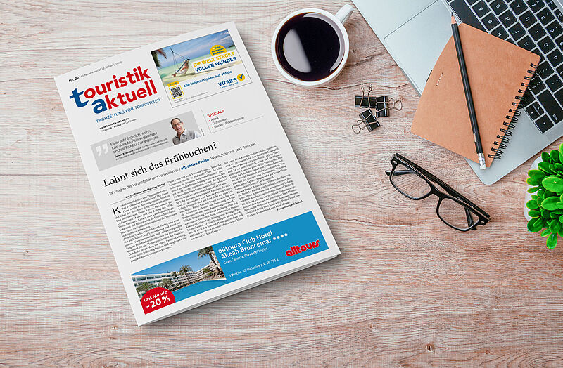 Auf einem Schreibtisch liegen eine Ausgabe der Fachzeitung touristik aktuell, eine Brille, ein Notizbuch mit Bleistift, eine Tasse Kaffee, Büroklammern, ein Laptop und eine kleine grüne Pflanze