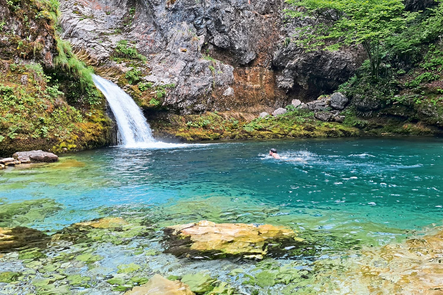 Kristallklarer Naturpool mit Wasserfall und Schwimmer in grüner Berglandschaft.