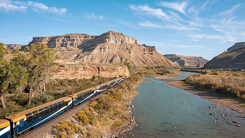 Ein blau-goldener Panoramazug fährt parallel zum Colorado River in den USA durch eine dramatische Canyon-Landschaft mit rötlichen Felswänden. Es herrscht sonniges Wetter, der Himmel ist blau mit einigen Schleierwolken.