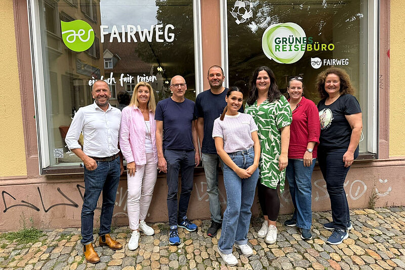 Eine Gruppe von acht Personen steht lächelnd vor einem Schaufenster mit den Logos Fairweg und Grünes Reisebüro. Die Menschen posieren gemeinsam auf einem gepflasterten Gehweg.