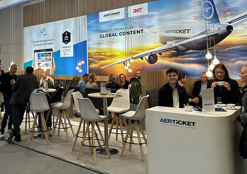 Messestand von Aerticket auf einer Tourismusmesse. Im Vordergrund ein Empfangstresen mit dem Aerticket-Logo und mehreren Personen, die sich unterhalten und Kaffee trinken. Dahinter befinden sich Stehtische mit weißen Barhockern sowie Sitzgruppen, an denen weitere Gespräche stattfinden. Im Hintergrund große Werbebanner von Aerticket und Giata mit einem Flugzeug und dem Slogan „Benefit from our global content“. Moderne Hängelampen und Pflanzen sorgen für eine einladende Atmosphäre.