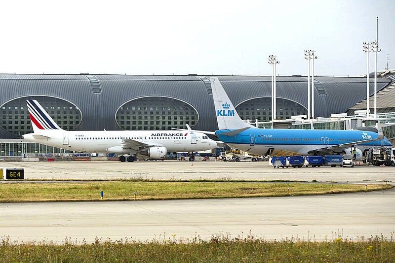 Zwei Flugzeuge Air France und KLM parken nebeneinander vor einem modernem Terminal mit geschwungener Architektur und Fluggastbrücken. Im Vordergrund ist ein Rollfeld zu sehen.