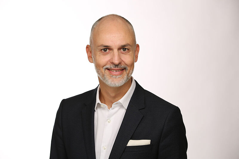 Ein lächelnder Manager mittleren Alters mit weißem Hemd und schwarzen Jackett vor einem Hintergrund mit weiß-grauem Farbverlauf.