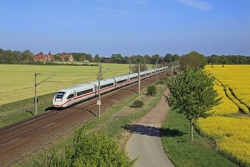 Ein moderner ICE-Zug der Deutschen Bahn fährt an einem sonnigen Tag durch eine ländliche Landschaft mit blühenden Rapsfeldern und grünen Wiesen. Links neben der Bahnstrecke ist ein großes Gebäude mit roten Dächern zu sehen. Der Himmel ist klar und blau, während eine kleine Landstraße parallel zur Bahnstrecke verläuft.