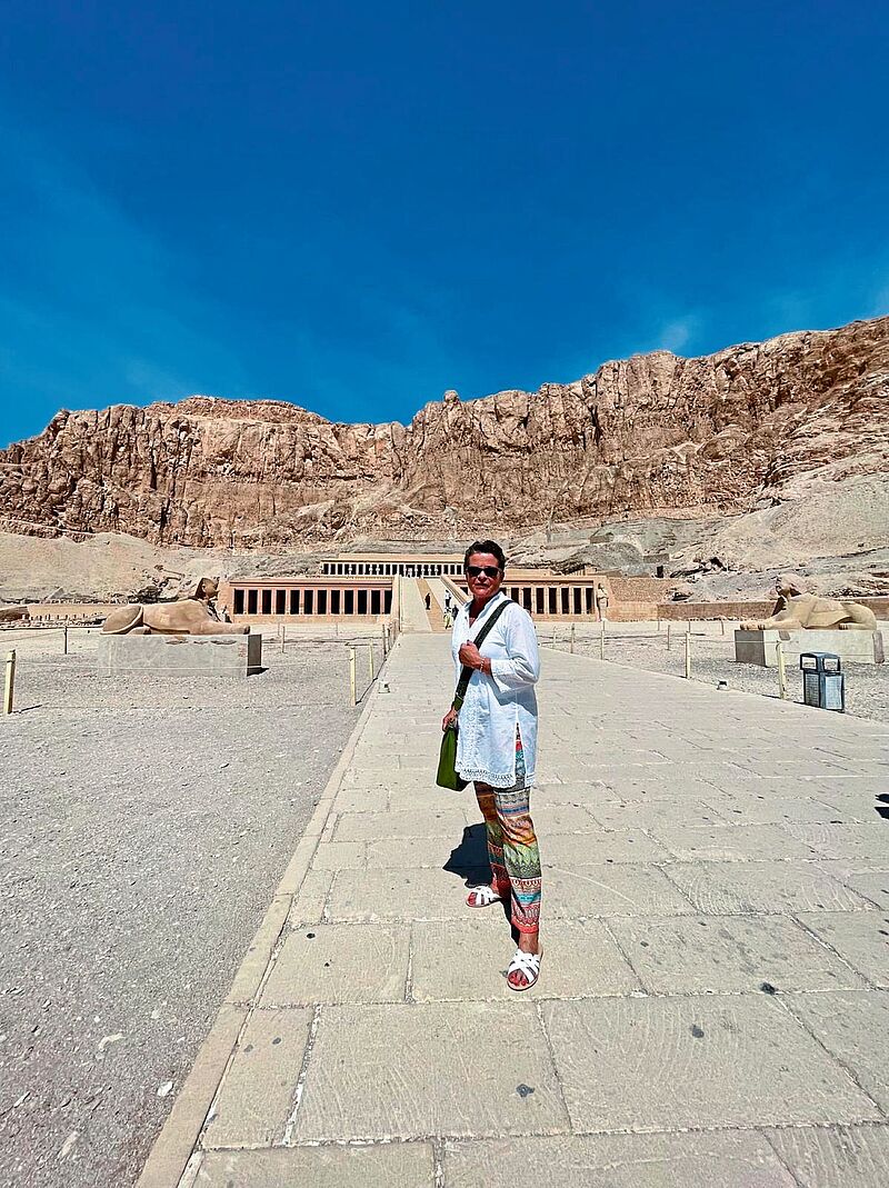 Terrassentempel der Hatschepsut in Luxor, Ägypten, umgeben von steilen Felsklippen unter klarem blauem Himmel. Davor steht eine Frau und lächelt in die Kamera