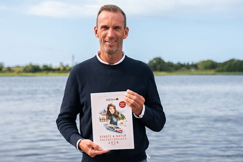 Der Vertriebschef des Flussreiseanbieters Arosa, Stefan Sprunk, steht mit dem neuen Katalog für 2026 in den Händen vor einem Fluss. Der Mittvierziger mit kurzem braunem Haar lächelt in die Kamera, er trägt einen dunkelblauen Pullover auf weißem Oberhemd.