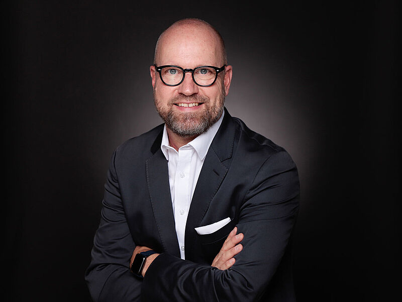 Ein Manager um die Fünfzig mit lichtem Haar und dicker Brille lächelt freundlich in die Kamera. Er trägt einen dunklen Anzug und ein weißes Oberhemd.