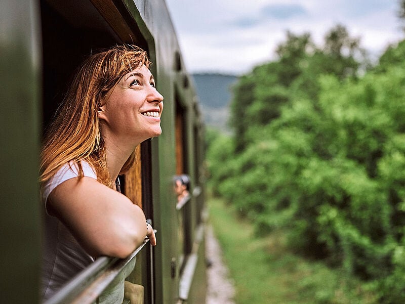Eine Lächelnde Frau mit rötlichen Haaren lehnt aus dem Fenster eines grünen Zuges und betrachtet eine vorbeiziehende grüne Waldlandschaft.
