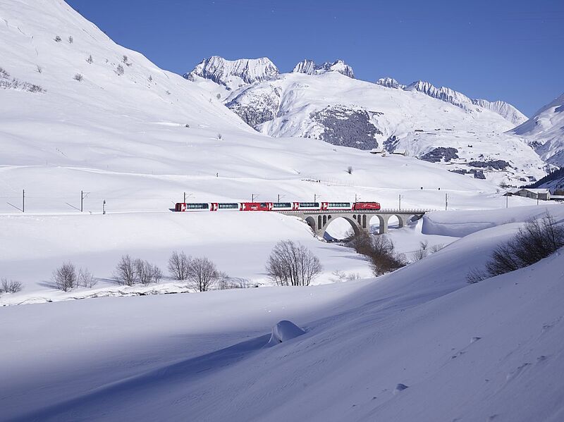 Ein roter Zug des Glacier Express fährt über ein steinernes Viadukt durch eine verschneite Berglandschaft in den Schweizer Alpen. Die Szenerie ist geprägt von tiefem Schnee, kahlen Bäumen und imposanten schneebedeckten Gipfeln unter klarem, blauem Himmel.