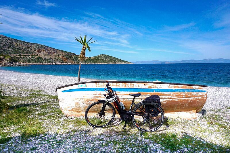 Ein schwarzes E-Bike steht vor einem alten, verwitterten Holzboot mit der Aufschrift „DIVA“ an einem weißen Kiesstrand. Im Hintergrund leuchten das türkisfarbene Meer und grüne Hügel unter einem strahlend blauen Himmel. Eine einzelne kleine Palmenpflanze wächst neben dem Boot und verleiht der Szenerie ein mediterranes Flair.