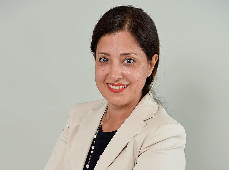 Porträtaufnahme einer Frau mit dunklen Haaren im cremefarbenen Blazer vor hellgrauem Hintergrund, mit freundlichem Lächeln und professioneller Ausstrahlung