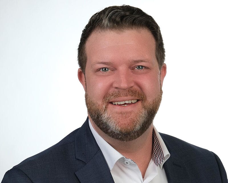 Porträt eines freundlich lächelnden Mannes im Business-Outfit vor hellem Hintergrund. Er trägt ein dunkelblaues Sakko mit Einstecktuch und ein weißes Hemd. Der Mann hat kurzes, dunkles Haar und einen gepflegten Bart.