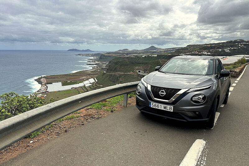 Panoramablick auf eine felsige Küstenlandschaft auf Gran Canaria bei wolkigem Wetter, mit dem Atlantik im Hintergrund und vereinzelten Palmen sowie einem Mietwagen im Vordergrund