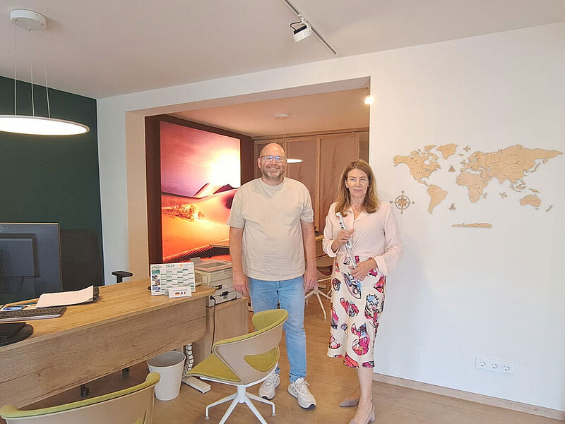 In einem modernen Reisebüro stehen eine Frau und ein Mann nebeneinander vor einer weißen Wand mit Holz-Weltkarte. Links im Vordergrund befindet sich ein Holzschreibtisch mit Computer, Kalender und Büromaterial, dahinter zwei grüne Bürostühle. Die Wand hinter dem Schreibtisch zeigt ein großes Landschaftsbild mit Reisterrassen und Bergen, im Nebenraum ist ein weiteres großes Fotomotiv mit Sanddünen zu sehen.