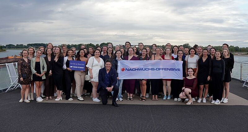 Eine große Gruppe junger Erwachsener posiert auf dem Deck eines Schiffs vor einem Flussufer. In der Mitte halten zwei Personen ein Banner mit der Aufschrift „NACHWUCHS-OFFENSIVE“. Eine andere Person hält ein Schild mit dem Logo von „Nicko Cruises“. Der Himmel ist bewölkt, und im Hintergrund ist die Landschaft mit Bäumen und Wasser zu sehen.